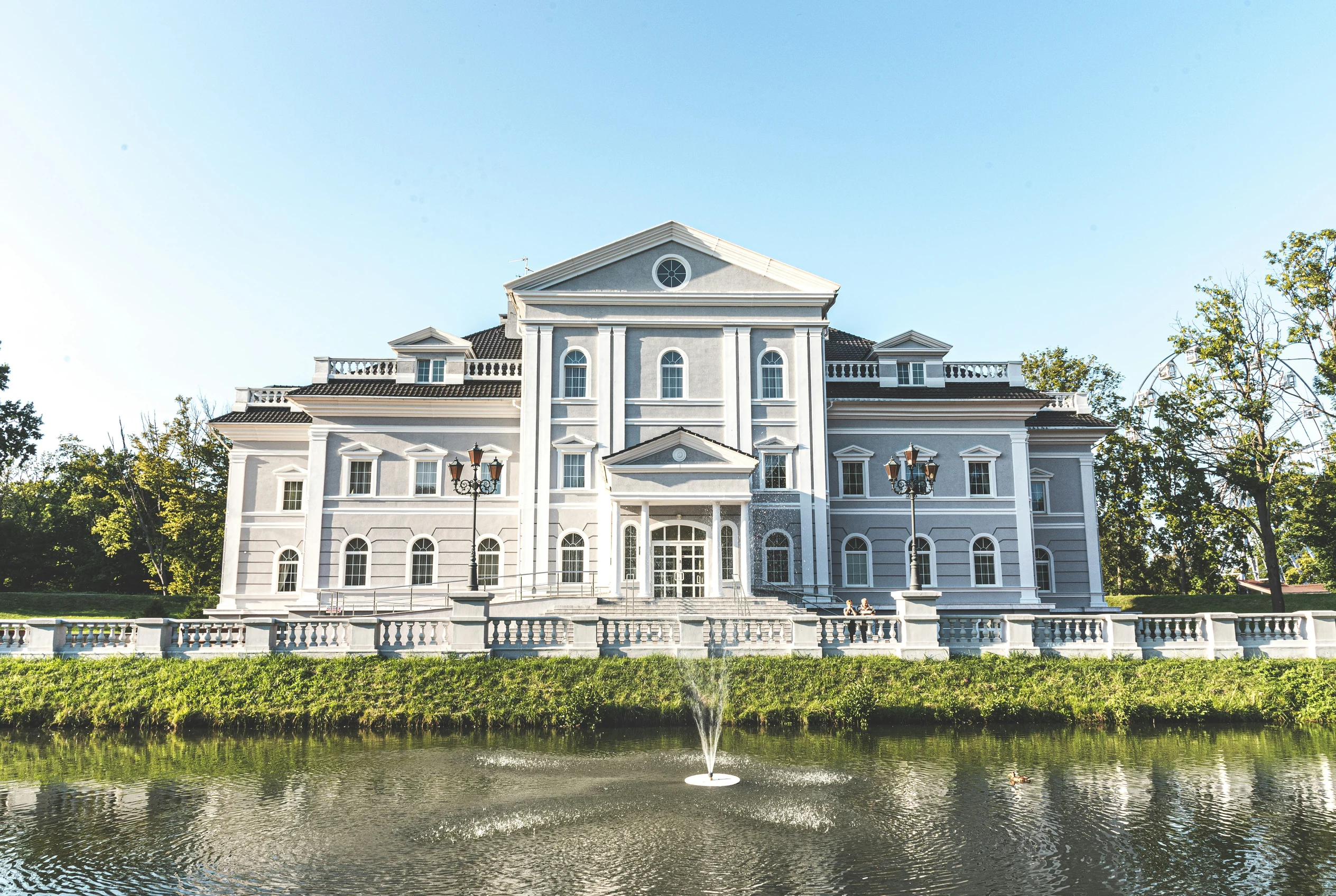 The finished building on the site: a three-storey mansion rendered in pale grey and white, with a pediment and round window over a columned entrance portico, arched windows, twin lamp posts, a stone balustrade along the top of the bank, and a fountain playing in the pond in front of it.