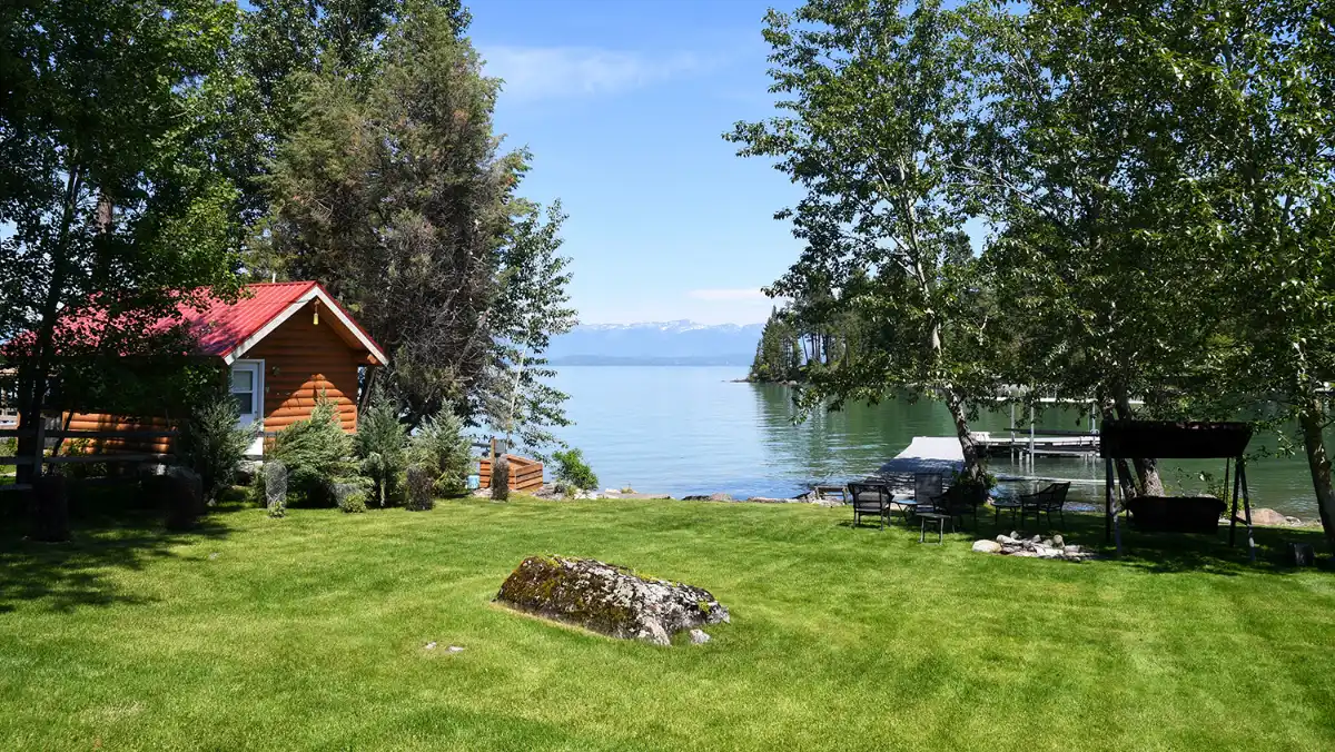 A mown lakeside lawn running down to calm water, with a small log cabin under mature birches and conifers, a timber dock to the right, and mountains on the far shore.